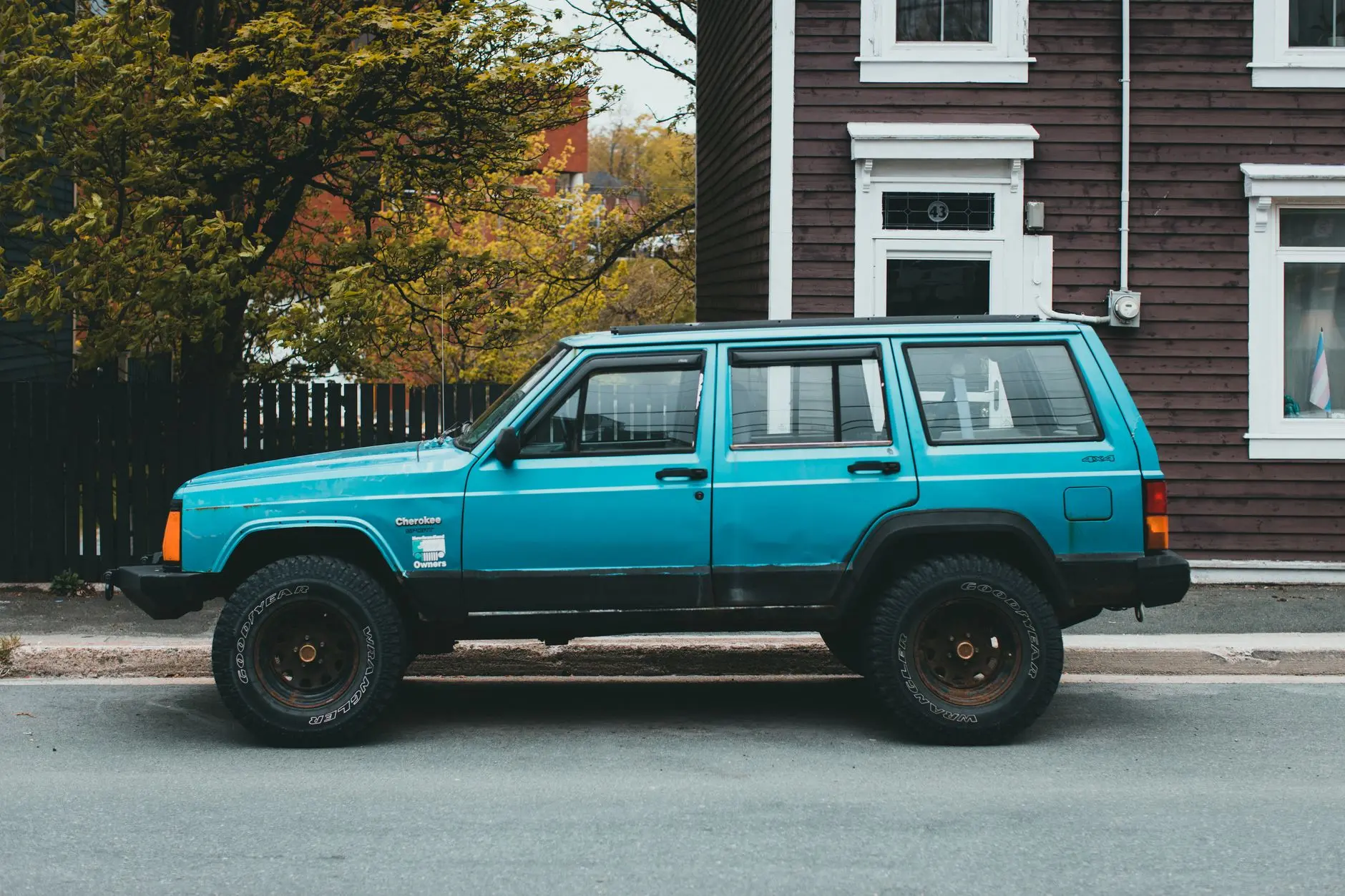 Bright blue vintage Jeep Cherokee parked by a rustic building on a tree-lined street.