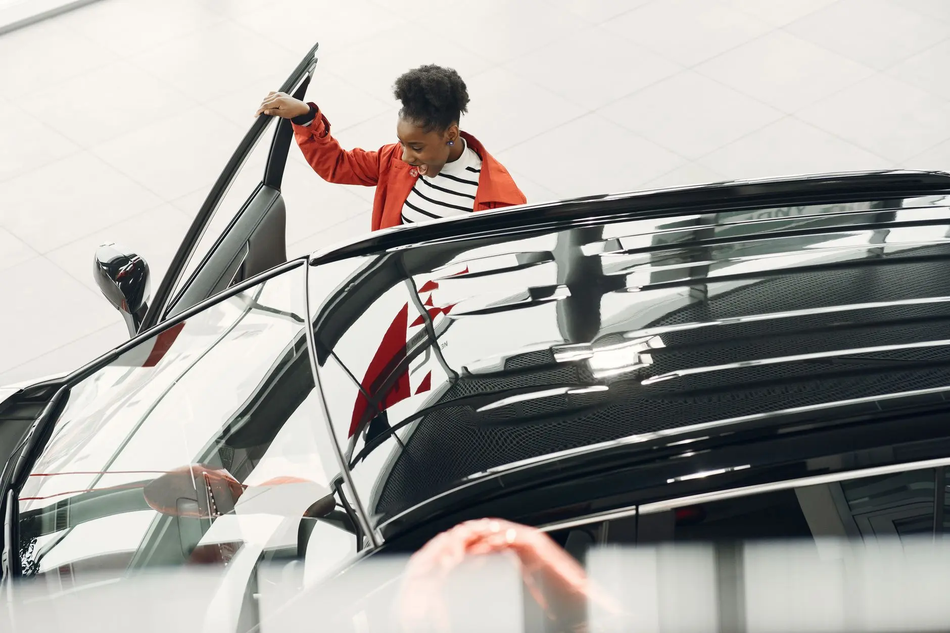 A woman in a red jacket opening a car door inside a modern car dealership.