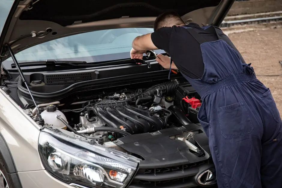 A mechanic in blue overalls works on a car engine in an outdoor setting.